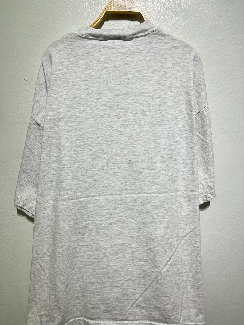 May include: A light gray, short-sleeved t-shirt hanging on a wooden hanger. The shirt has a mock neck and a simple, minimalist design. The fabric appears to be a soft, comfortable material.