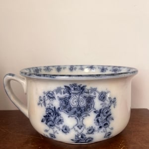 Op de afbeelding: Een witte keramische kamerpot met een blauw bloemenpatroon in transferdruk. De pot heeft één handvat aan de zijkant en een brede, afgeronde rand die is versierd met een herhalend blauw bloemmotief langs de gehele bovenste rand.