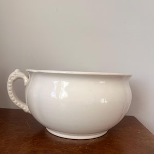 Puede incluir: Un orinal de cerámica blanca con un cuenco liso y redondeado, y un asa lateral única que presenta una textura decorativa de cuentas a lo largo de su borde exterior. El recipiente tiene una parte superior ancha y abierta y una base circular.