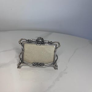 Peut inclure: Un petit cadre photo rectangulaire en métal argenté avec un motif floral en relief orné au centre, en haut et en bas. Le cadre présente des bords décoratifs incurvés et contient un insert rectangulaire uni de couleur claire.
