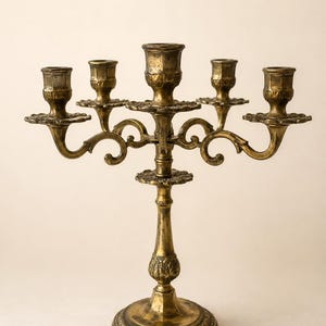 May include: A five-arm candelabra crafted from aged brass with a weathered gold finish. The piece features a central stem with a circular base, ornate floral detailing at the joints, and curved arms supporting five individual candle cups.