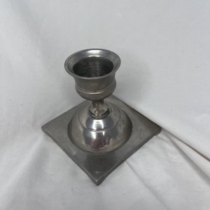 Könnte beinhalten: Ein silberfarbener Kerzenhalter mit quadratischem Sockel. Der Halter hat eine runde, ausgestellte Oberseite für eine Kerze. Der Sockel ist quadratisch mit einem rautenförmigen Design. Der Kerzenhalter ist aus Metall.