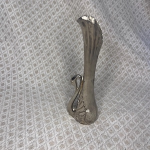 Pode incluir: Vaso prateado em forma de cisne. O vaso tem um pescoço longo e fino com uma abertura alargada e uma cabeça e pescoço de cisne formando a base. O vaso tem um design texturizado e canelado no pescoço e no corpo.