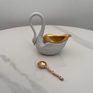 May include: A white swan-shaped dish with a textured feather pattern and a smooth gold-colored interior, accompanied by a small gold-colored spoon featuring a textured, branch-like handle with small nodes along its length.