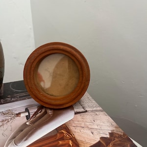 May include: A small, circular wooden picture frame with a smooth, medium-brown finish. The frame features a simple, rounded profile and contains a plain, light-coloured insert, presenting a clean and straightforward design for displaying small images.