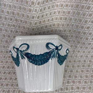 May include: White ceramic planter with teal bow and garland design. The planter has a ribbed texture and a hexagonal shape. Decorative accent for home decor.