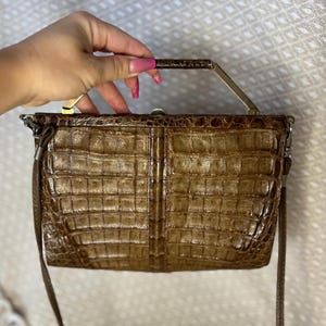 May include: A brown, textured handbag with a rectangular shape and a metal handle. The bag has a crocodile skin pattern and a long, thin strap. The handle is a gold color.