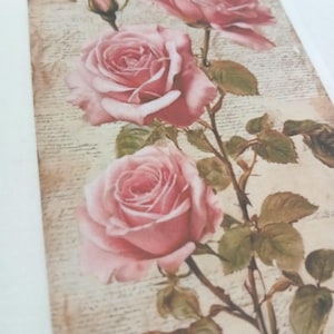 May include: A rectangular print featuring pink roses with green leaves and thorny stems against a vintage, cream-colored background. The roses are in full bloom, with a bud visible. The background has faint script.