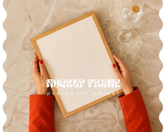 May include: A wooden picture frame with a white interior is held by a person wearing a red blazer. The frame is on a marble surface, with a small glass bottle and a small glass cup in the background. The text "MOCKUP FRAME" is visible.