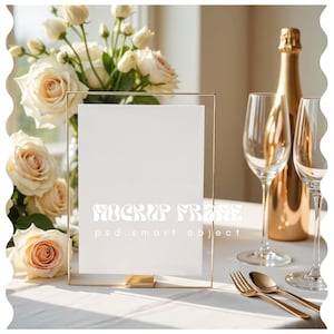 May include: A gold-framed clear glass sign holder with a white insert, set on a white tablecloth. Champagne glasses, a gold bottle, and gold cutlery are arranged nearby. A bouquet of cream-colored roses is in the background.