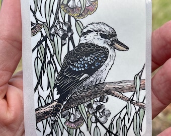 May include: A black and white illustration of a kookaburra perched on a branch with green leaves and pink flowers. The bird has blue feathers on its back and a long beak.