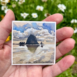 Könnte beinhalten: Ein quadratischer Aufkleber mit einer Aquarell-Landschaft, die einen dunklen Felsen im Meer zeigt, der sich im nassen Sand unter einem blau-weißen, bewölkten Himmel spiegelt. Der Text carrie carlson art steht unten rechts in Schwarz.