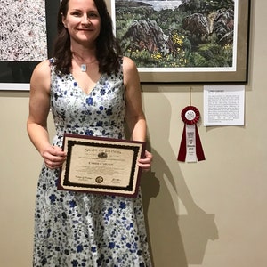 May include: A woman in a floral dress holds a certificate of recognition from the State of Illinois. The certificate is for Carrie Carlson. The woman is standing in front of a wall with two framed pieces of art. The art on the right is a landscape with a red ribbon.