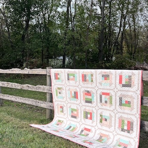 Könnte beinhalten: Eine handgefertigte Steppdecke mit Log Cabin-Design, drapiert über einem Holzzaun. Die Steppdecke zeigt quadratische Blöcke mit verschiedenen Farben, darunter Koralle, Grün und Grau. Der Hintergrund ist eine natürliche Umgebung mit Bäumen und Gras.