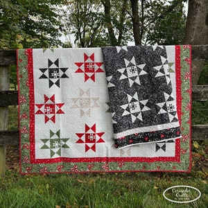 May include: Two quilts hanging on a wooden fence. One quilt is white with red, black, and green star patterns. The other quilt is black and white with star patterns. The quilts have red and green borders.