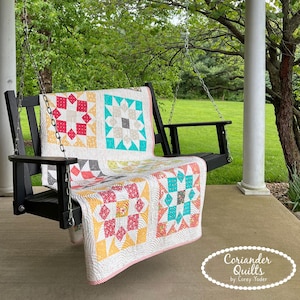 May include: A colourful quilt with star patterns in red, turquoise, and yellow is draped over a black wooden porch swing. The quilt has a white background and is hanging outside on a porch. The quilt is made by Coriander Quilts.
