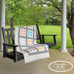 May include: A black wooden porch swing with a colourful quilt draped over it. The quilt features a grid pattern with squares of various colours, including red, orange, and blue. The swing is hanging from chains on a porch with white columns, with green trees in the background.