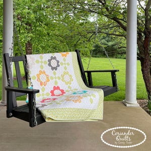 Op de afbeelding: Een zwarte houten veranda schommel met een bloemenquilt eroverheen. De quilt heeft kleurrijke bloemen op een witte achtergrond, met een groene rand. De schommel hangt aan kettingen aan een overdekte veranda.