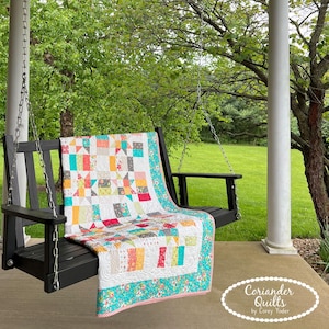May include: A patchwork quilt in shades of red, teal, yellow, and grey is draped over a black porch swing. The quilt has a variety of square and rectangular patterns. The border features a floral design. The text "Coriander Quilts by Corey Yoder" is visible.