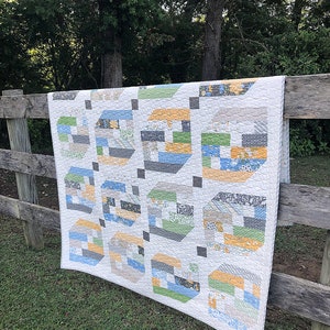 May include: A handmade quilt with a repeating geometric pattern of colorful blocks in shades of blue, green, gray, and yellow. The quilt is draped over a wooden fence, with a background of green grass and trees.
