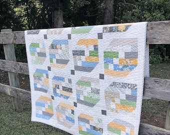 May include: A handmade quilt with a repeating geometric pattern of colorful blocks in shades of blue, green, gray, and yellow. The quilt is draped over a wooden fence, with a background of green grass and trees.