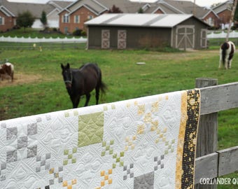May include: A handmade quilt with a white background, featuring a geometric pattern in shades of gray, green, and gold. The quilt is draped over a wooden fence in front of a grassy field with horses grazing in the background.