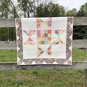 May include: A handmade quilt with a star pattern, featuring a white background with a scalloped design. The star is composed of colorful floral and geometric fabrics. The quilt is bordered with a dark floral fabric and a pink edge.