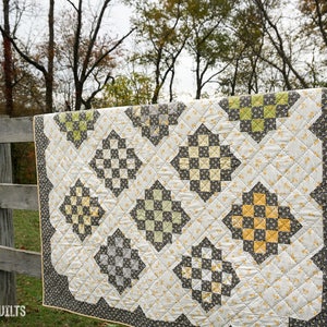 May include: A handmade quilt with a repeating diamond pattern in shades of gray, yellow, and green, hanging on a wooden fence. The quilt has a scalloped edge and is made of quilted fabric. The text "coriander quilts" is visible.