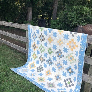 Op de afbeelding: Een handgemaakte quilt met een witte achtergrond en een herhalend patroon van blauwe sterren en geometrische blokken in grijstinten, groen en geel. De quilt heeft een lichtblauwe rand en is over een houten hek gedrapeerd.