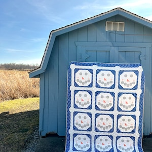 May include: A handmade quilt featuring a repeating pattern of snowman faces. The quilt has a white background with blue polka dots and a blue border. The snowman faces have gray eyes, orange noses, and pink smiles. The quilt is displayed against a blue shed.