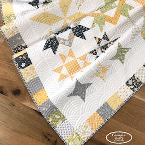 May include: A handmade quilt featuring star patterns in yellow, gray, and black on a white background. The quilt has a scalloped edge and is made of various floral and geometric patterned fabrics. The quilt is placed on a wooden surface.