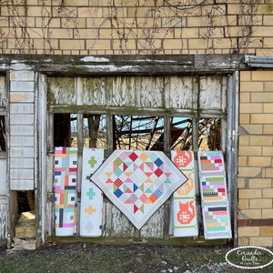 Op de afbeelding: Een display van handgemaakte quilts met diverse geometrische patronen. De quilts tonen ontwerpen zoals vierkanten, kruisen en een kattenmotief. De kleuren zijn rood, oranje, blauw, groen en wit. De quilts hangen in een oude houten raamkozijn.