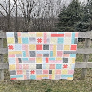 May include: A colorful patchwork quilt with a geometric design, featuring squares and rectangles in shades of pink, yellow, gray, and teal. The quilt is adorned with coral-colored star accents and hangs over a wooden fence.