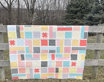 Può includere: Una colorata trapunta patchwork con un design geometrico, con quadrati e rettangoli nei toni del rosa, giallo, grigio e verde acqua. La trapunta è adornata da accenti a stella color corallo ed è appesa a una staccionata di legno.