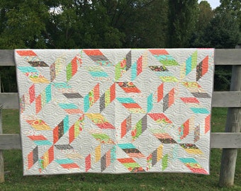 May include: A handmade quilt with a geometric pattern of colorful rectangles in shades of coral, teal, green, and gray. The quilt is displayed outdoors, draped over a wooden fence, with a background of green grass and trees.