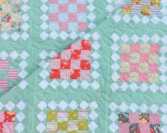 Puede incluir: Un edredón hecho a mano con un fondo verde menta y un patrón repetido de diamantes blancos. El edredón presenta bloques cuadrados de varias telas estampadas en diseños rojos, verdes y florales. El edredón está acolchado a máquina con un patrón de vieira.