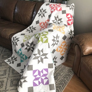 May include: A white quilt with a star pattern in various colors, including gray, red, orange, green, teal, and purple. The quilt is draped over a brown leather couch. The quilt has a quilted design and the words "coriander quilts" are visible.