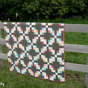 Op de afbeelding: Een handgemaakte quilt met een donkergrijze achtergrond en een herhalend patroon van kleurrijke vierkanten. De vierkanten zijn in tinten rood, wit, blauw, geel en groen. De quilt hangt aan een houten hek in een grasveld.