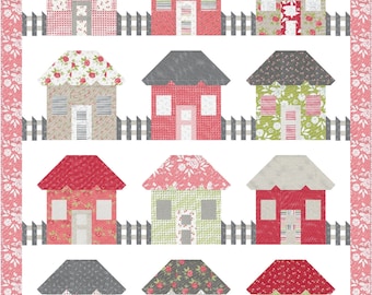Puede incluir: Un edredón de retazos con doce casas estilizadas con tejados grises, rojos y verdes. Las casas tienen varios diseños florales y estampados. Una valla de estacas gris corre frente a las casas. El edredón está bordeado con un estampado floral.