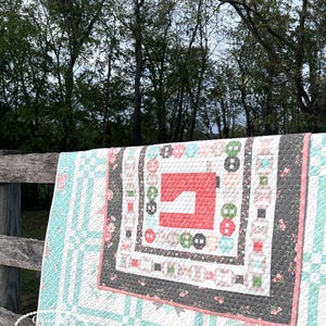 May include: A patchwork quilt with a coral sewing machine motif at its centre. The quilt has a white base with a grid design, teal stripes, floral patterns, and a dark grey border. It is draped over a wooden fence, with trees in the background.