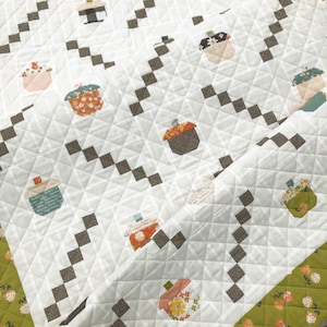 May include: White quilted blanket with a repeating pattern of colorful jar-shaped designs. The jars feature various patterns and colors, including floral and geometric prints. Brown square accents and a green floral border complete the design.