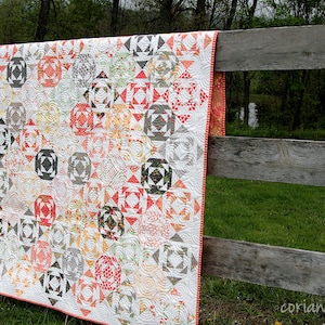May include: A handmade quilt featuring a repeating geometric pattern in red, grey, orange, and green on a white background. The quilt is draped over a weathered wooden fence. The border is red and white striped.