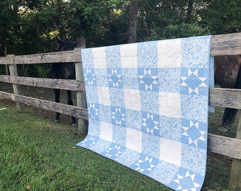 Könnte beinhalten: Eine blau-weiße Steppdecke mit Stern- und Blumenmuster ist über einen Holzzaun drapiert. Die Steppdecke hat ein Rasterdesign mit abwechselnden Quadraten aus gemustertem Stoff. Der Hintergrund umfasst ein Pferd und grünes Laub.