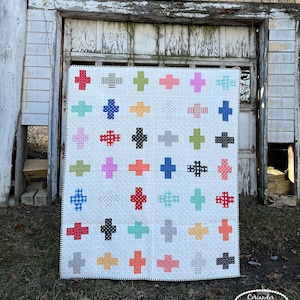 Może przedstawiać: Biała kołdra z powtarzającym się wzorem krzyża w różnych kolorach i wzorach, w tym paski, kropki i krata. Kołdra ma czarno-białą obwódkę w paski i jest prezentowana na zewnątrz. Wykonane przez Coriander Quilts.