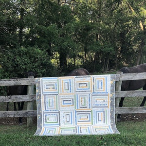 May include: A rectangular quilt with a log cabin design, featuring squares of various sizes and colors. The quilt's color palette includes white, blue, green, and yellow. The quilt is displayed outdoors, draped over a wooden fence.