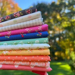 May include: A stack of folded fabric squares featuring various patterns including cherries, flowers, gingham, and polka dots in colors of dark gray, white, pink, blue, teal, yellow, and orange, arranged in a vertical column.