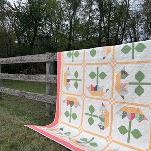 May include: A handmade quilt with a floral and bird pattern, displayed outdoors. The quilt features a white background with colorful squares, each containing a flower or bird design. The border is a coral color, and the quilt is draped over a wooden fence.