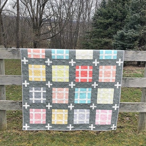 May include: A handmade quilt with a gray background and colorful squares. Each square features a different pastel color and a white cross design. The quilt is displayed outdoors, draped over a wooden fence.