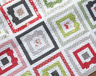 May include: Close-up of a quilt with a repeating square pattern. The squares feature concentric rectangles in red, green, gray, and black, with white accents. The quilt has a patchwork design with various patterned fabrics.