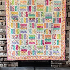 Op de afbeelding: Een rechthoekige quilt met een patchworkontwerp van veelkleurige stoffen rechthoeken in tinten roze, geel, oranje, groen en paars. De quilt heeft een rand met bloemenpatroon en is afgewerkt met een felroze bies langs de rand.